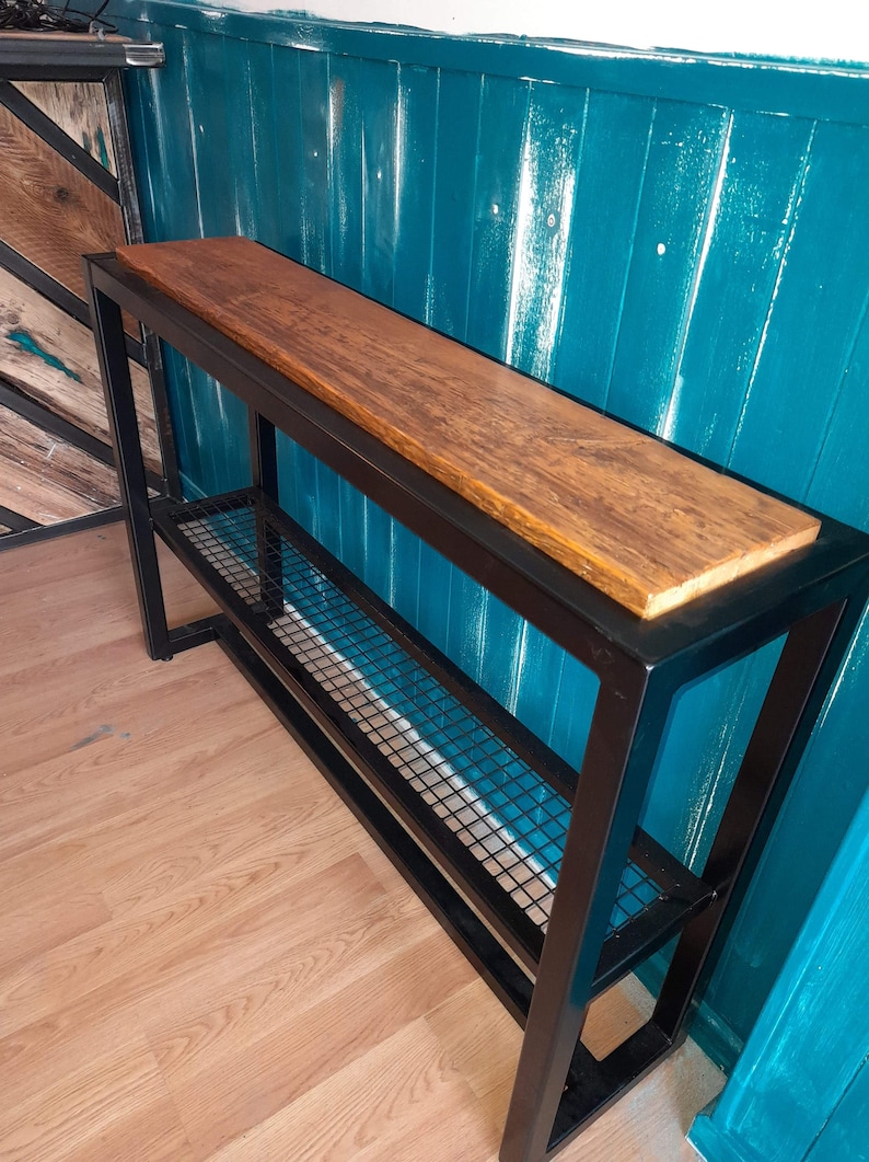 May include: A black metal console table with a wooden top and a wire shelf. The table is against a teal wall.