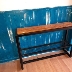 May include: A black metal console table with a wooden top and a wire shelf. The table is against a teal blue wall with white painted wood paneling.