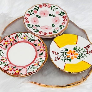 Könnte beinhalten: Drei kleine, dekorative Keramikplatten mit gewellten Rändern. Jede Platte hat ein einzigartiges, handgemaltes Blumendesign in Rosa, Gelb und Rot mit goldenem Rand. Die Teller sind auf einem grau-goldenen Dekotablett angeordnet.