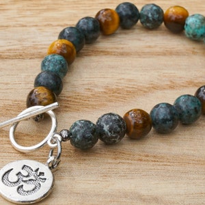 May include: A bracelet made of round, polished beads in teal, brown, and gold. It has a silver toggle clasp and a charm with the Om symbol. The bracelet is presented on a wooden surface. The beads are approximately 0.8 cm in diameter.