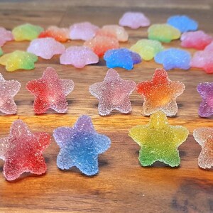 Puede incluir: Una colecci&oacute;n de caramelos coloridos en forma de estrella con una textura azucarada. Los caramelos muestran un degradado de colores, incluyendo rojo, naranja, amarillo, verde, azul y morado. Algunos caramelos tienen forma de nube. Los caramelos est&aacute;n dispuestos sobre una superficie de madera.