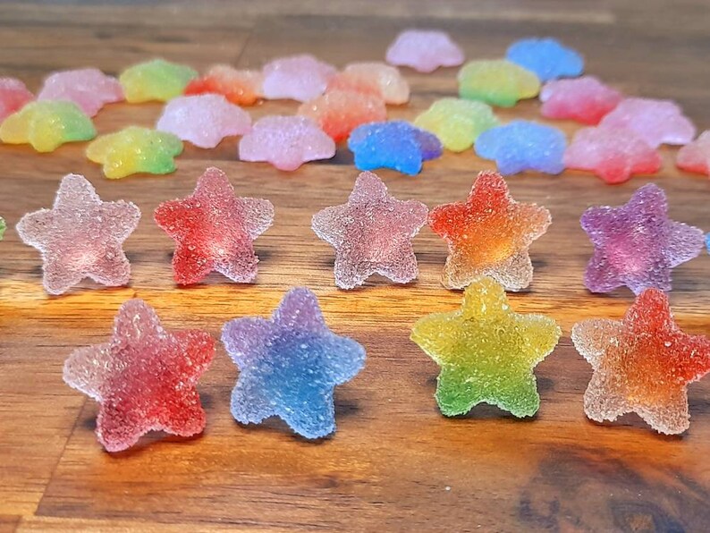 Puede incluir: Una colecci&oacute;n de caramelos coloridos en forma de estrella con una textura azucarada. Los caramelos muestran un degradado de colores, incluyendo rojo, naranja, amarillo, verde, azul y morado. Algunos caramelos tienen forma de nube. Los caramelos est&aacute;n dispuestos sobre una superficie de madera.
