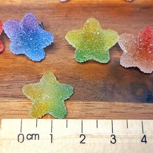 Puede incluir: Primer plano de caramelos con forma de estrella y coloridos con una capa de az&uacute;car. Los caramelos son de color rojo, azul, verde y amarillo. Una regla se coloca junto a los caramelos, midiendo aproximadamente 2.5 cm de largo.