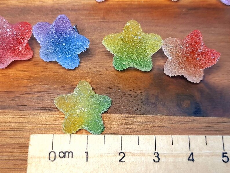 Puede incluir: Primer plano de caramelos con forma de estrella y coloridos con una capa de az&uacute;car. Los caramelos son de color rojo, azul, verde y amarillo. Una regla se coloca junto a los caramelos, midiendo aproximadamente 2.5 cm de largo.