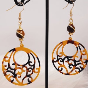 Pode incluir: Um par de brincos pendurados com um padrão de tartaruga. Cada brinco tem um gancho dourado, uma conta marrom enrolada em fio dourado e um pingente circular com um design vazado. Os brincos são exibidos em um suporte preto.
