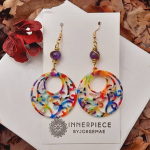 Pode incluir: Brincos dourados com um design circular vazado em cores do arco-íris. Cada brinco apresenta uma conta roxa e o texto "INNERPIECE BY JORGEMAE" em um cartão branco.