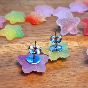 Puede incluir: Pendientes de bot&oacute;n en forma de estrella con cierre de poste. Los pendientes est&aacute;n hechos de un material transl&uacute;cido y colorido, con un degradado de colores. Otros objetos en forma de estrella est&aacute;n dispersos sobre una superficie de madera.