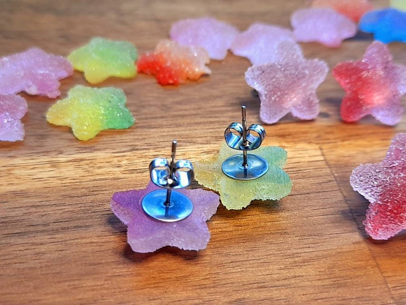 Puede incluir: Pendientes de bot&oacute;n en forma de estrella con cierre de poste. Los pendientes est&aacute;n hechos de un material transl&uacute;cido y colorido, con un degradado de colores. Otros objetos en forma de estrella est&aacute;n dispersos sobre una superficie de madera.