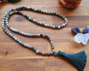 Könnte beinhalten: Eine Perlen-Mala-Kette mit einer tealfarbenen Quaste. Die Perlen sind eine Mischung aus Farben, darunter Grün, Weiß und Lila. Die Kette wird auf einer Holzoberfläche mit anderen dekorativen Gegenständen präsentiert.