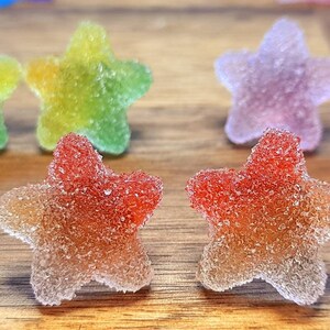 Puede incluir: Primer plano de caramelos de goma en forma de estrella con una capa de az&uacute;car. Los caramelos tienen un degradado de colores, incluyendo rojo, naranja, amarillo, verde y morado. Los caramelos est&aacute;n sobre una superficie de madera.