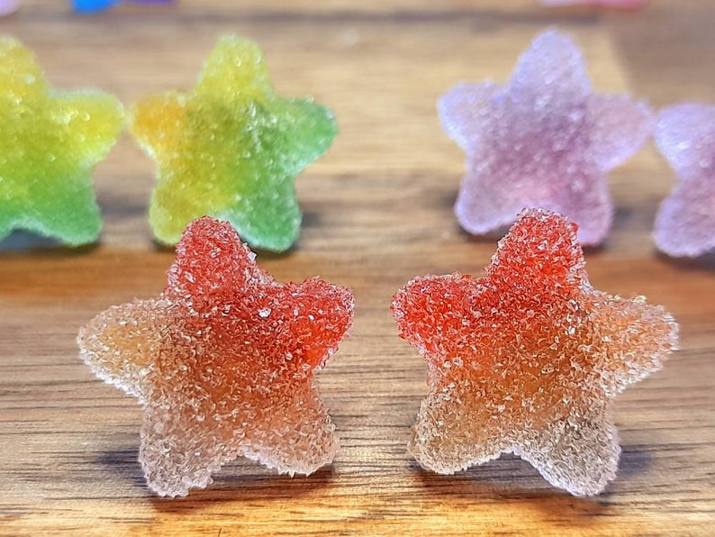 Puede incluir: Primer plano de caramelos de goma en forma de estrella con una capa de az&uacute;car. Los caramelos tienen un degradado de colores, incluyendo rojo, naranja, amarillo, verde y morado. Los caramelos est&aacute;n sobre una superficie de madera.