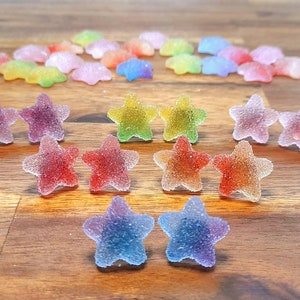 Puede incluir: Un surtido de caramelos coloridos en forma de estrella con una capa de az&uacute;car. Los caramelos son en tonos de rojo, naranja, amarillo, verde, morado y azul. Algunos est&aacute;n dispuestos en pares, otros est&aacute;n dispersos.