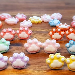 Op de afbeelding: Een verzameling kleurrijke snoepjes in de vorm van poten in verschillende kleuren, waaronder roze, oranje, geel, blauw, groen en paars. Elk snoepje heeft een witte pootafdruk in het midden, op een houten achtergrond.