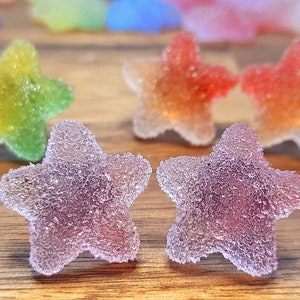 Puede incluir: Primer plano de caramelos coloridos en forma de estrella con una capa de az&uacute;car. Los caramelos son de color p&uacute;rpura, rojo, naranja, amarillo y verde. Los caramelos est&aacute;n sobre una superficie de madera.