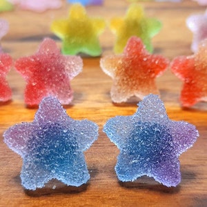 Puede incluir: Primer plano de caramelos coloridos en forma de estrella con una capa de az&uacute;car. Los caramelos son de varios colores, incluyendo rojo, naranja, amarillo, verde y azul. Est&aacute;n dispuestos sobre una superficie de madera.