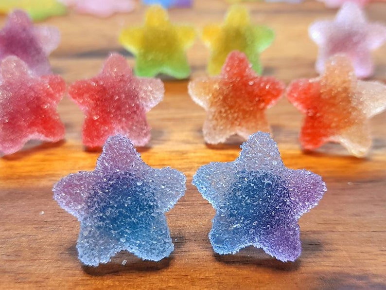 Puede incluir: Primer plano de caramelos coloridos en forma de estrella con una capa de az&uacute;car. Los caramelos son de varios colores, incluyendo rojo, naranja, amarillo, verde y azul. Est&aacute;n dispuestos sobre una superficie de madera.