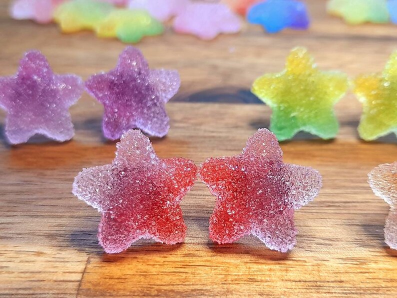 Puede incluir: Primer plano de caramelos con forma de estrella coloridos con una capa de az&uacute;car. Los caramelos son de color rojo, morado, amarillo y verde, dispuestos sobre una superficie de madera. Los caramelos tienen un aspecto transl&uacute;cido.