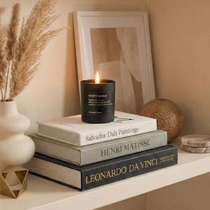 May include: A black cylindrical candle with a lit flame sits atop a stack of three books titled Salvador Dali Paintings, Henri Matisse, and Leonardo da Vinci. The candle label reads Buvcte Saidle. The books are arranged on a flat surface.