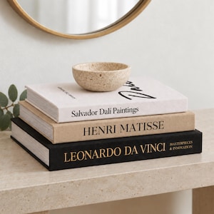 May include: A small, beige, textured stone bowl sits atop a stack of three hardcover books. The top book is white with black text reading Salvador Dali Paintings, the middle is tan with Henri Matisse, and the bottom is black with gold text.