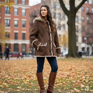May include: A brown suede coat with a faux fur-lined hood and toggle closures. The coat has cream-colored trim and is worn with dark blue jeans and brown knee-high boots. The background shows a park with fallen leaves and buildings.