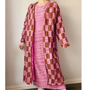 May include: A long, pink dress is paired with a long, open, brown and pink checkered coat. The coat has a checkerboard pattern in shades of brown and pink. The dress is a solid pink color. The outfit is worn by a person.