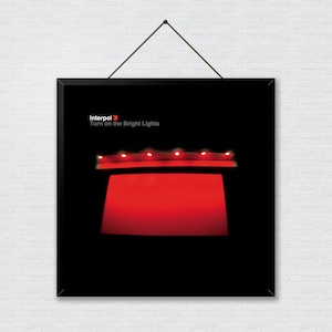 Può includere: Copertina dell'album in bianco e nero con il nome della band "Interpol" e il titolo dell'album "Turn on the Bright Lights" in testo bianco. La copertina dell'album presenta un palco rosso con cinque luci rosse sopra.