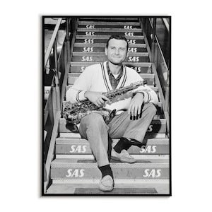 Puede incluir: Fotografía en blanco y negro enmarcada de un hombre sentado en unas escaleras exteriores, sosteniendo un saxofón. Las escaleras tienen las letras "SAS" impresas. El hombre lleva un suéter, vaqueros y mocasines.