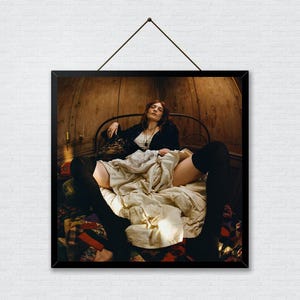 Puede incluir: Una fotografía enmarcada muestra a una persona reclinada en una cama, vistiendo una chaqueta negra y medias negras hasta el muslo. La cama está cubierta con una manta color crema y un edredón estampado. El fondo presenta paneles de madera.