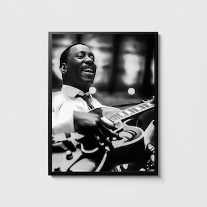 Puede incluir: Fotografía enmarcada en blanco y negro de un músico sonriente tocando una guitarra eléctrica. El músico lleva camisa blanca y corbata. La guitarra tiene un cuerpo curvo y varias cuerdas.