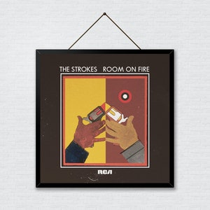 May include: Framed album art for The Strokes' "Room on Fire." The artwork features a split background of yellow and maroon, with two hands holding lighters. The band's name and album title are at the top.