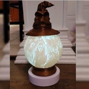 May include: A 3D printed lamp shaped like the Sorting Hat from the Harry Potter series. The lamp is lit and the light shines through a translucent sphere with an image of characters from the Harry Potter series.