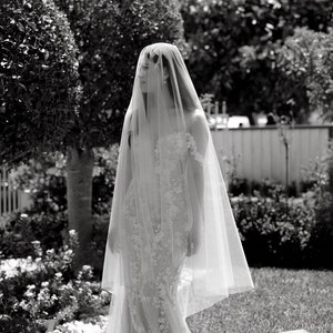 May include: A bride wearing a white wedding dress with a long veil. She is standing in a garden with green bushes and grass.