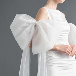 May include: A white satin mini dress with a large white tulle bow on one shoulder. The dress has a fitted bodice and a slightly flared skirt.