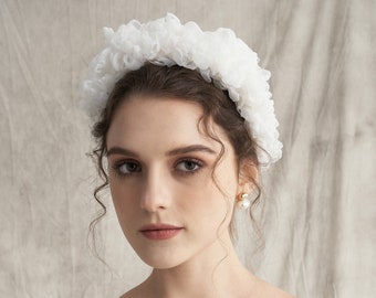 May include: A woman wearing a white, ruffled headband. She is also wearing gold and pearl earrings and a white strapless top. The background is a neutral, textured fabric.