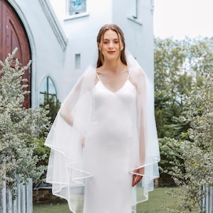 May include: A woman wearing a white satin wedding dress with a long veil. She is standing in front of a white building with a wooden door.