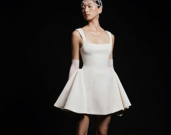 May include: A white sleeveless mini dress with a square neckline and a flared skirt. The dress is worn with white gloves and silver shoes with large bows.