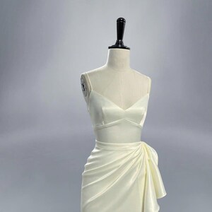 May include: A white satin dress with a fitted bodice and a flowing skirt. The skirt has a draped design and a bow detail on the side.