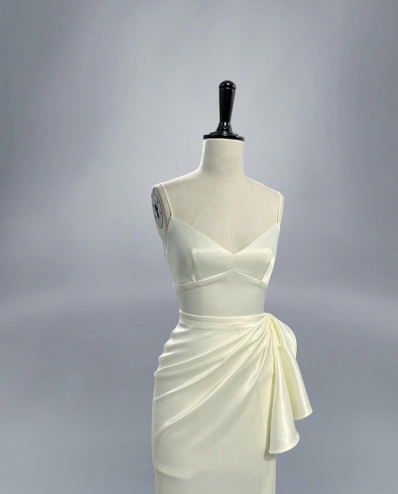 May include: A white satin dress with a fitted bodice and a flowing skirt. The skirt has a draped design and a bow detail on the side.