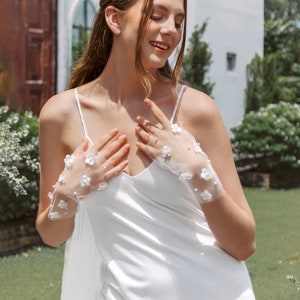 May include: A woman wearing a white satin slip dress with a white lace overlay and white floral lace gloves. She is standing in front of a white building with a wooden door.