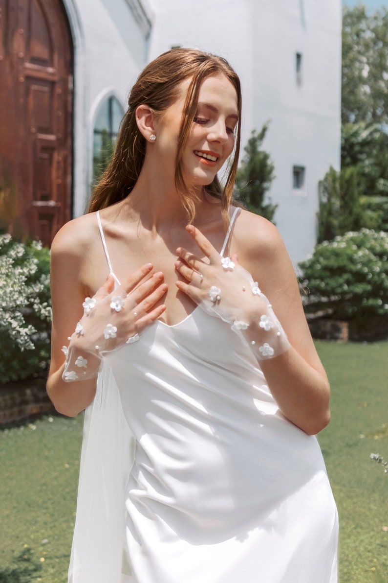 May include: A woman wearing a white satin slip dress with a white lace overlay and white floral lace gloves. She is standing in front of a white building with a wooden door.