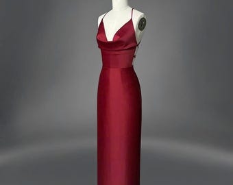 Könnte beinhalten: Ein bodenlanges, bordeauxfarbenes Abendkleid auf einer Schaufensterpuppe. Das Kleid hat einen Wasserfallausschnitt, Spaghettiträger und ein figurbetontes Oberteil, das in einen geraden Rock übergeht. Der Rücken des Kleides hat ein zartes, offenes Design.