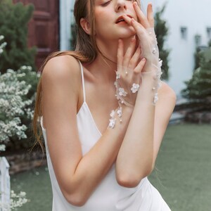 May include: A woman wearing a white slip dress with delicate floral lace gloves. The gloves have small white flowers and pearl accents.