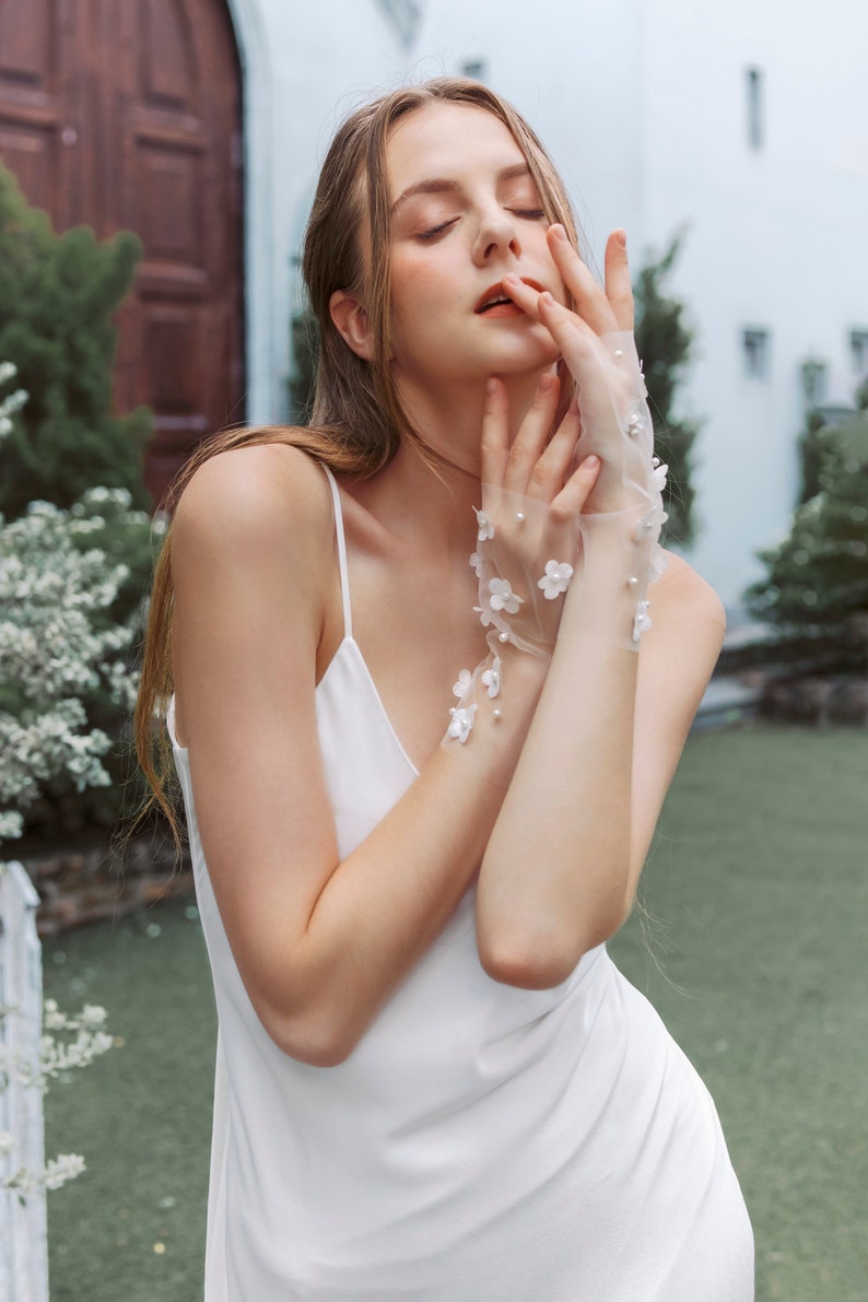 May include: A woman wearing a white slip dress with delicate floral lace gloves. The gloves have small white flowers and pearl accents.