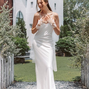 May include: A woman wearing a white satin dress with a lace overlay and a veil. She is standing in front of a white building with green grass and a wooden fence.