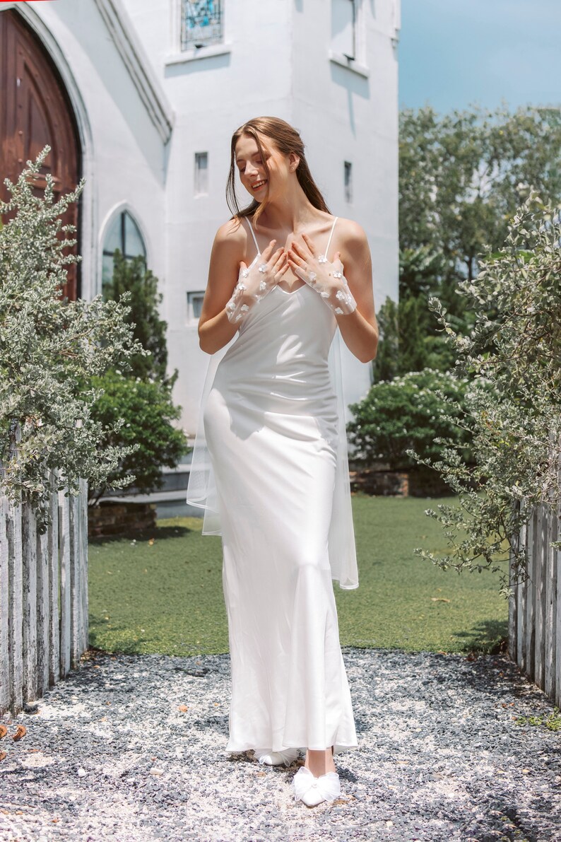 May include: A woman wearing a white satin dress with a lace overlay and a veil. She is standing in front of a white building with green grass and a wooden fence.