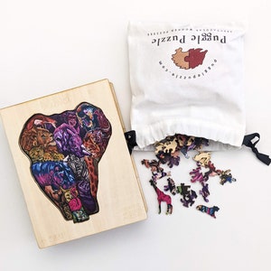 Puede incluir: Una caja de rompecabezas de madera con una imagen colorida de animales en forma de elefante. Las piezas del rompecabezas est&aacute;n esparcidas sobre una superficie blanca junto a una bolsa de tela blanca con el texto "Puzzle Puzzle" impreso en ella.