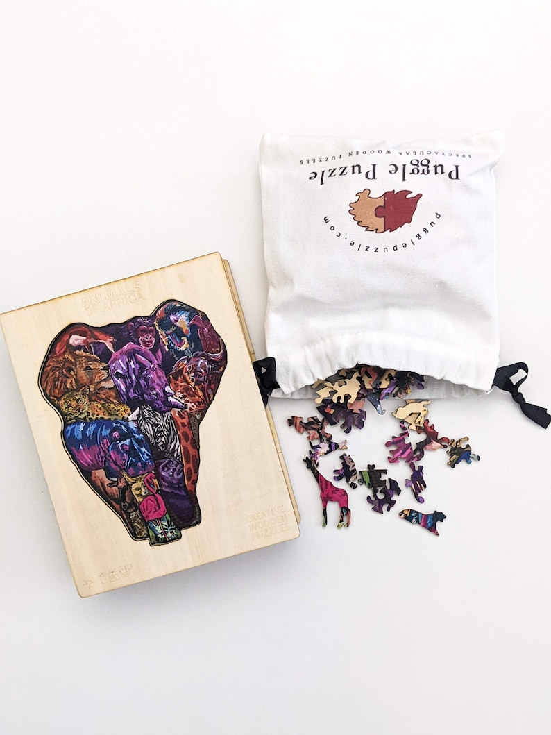Puede incluir: Una caja de rompecabezas de madera con una imagen colorida de animales en forma de elefante. Las piezas del rompecabezas est&aacute;n esparcidas sobre una superficie blanca junto a una bolsa de tela blanca con el texto "Puzzle Puzzle" impreso en ella.