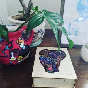 Puede incluir: Una caja de rompecabezas de madera con un dise&ntilde;o de elefante colorido. La caja est&aacute; abierta y las piezas del rompecabezas son visibles. La caja est&aacute; sobre una mesa de madera con una planta y un marco de imagen en el fondo.