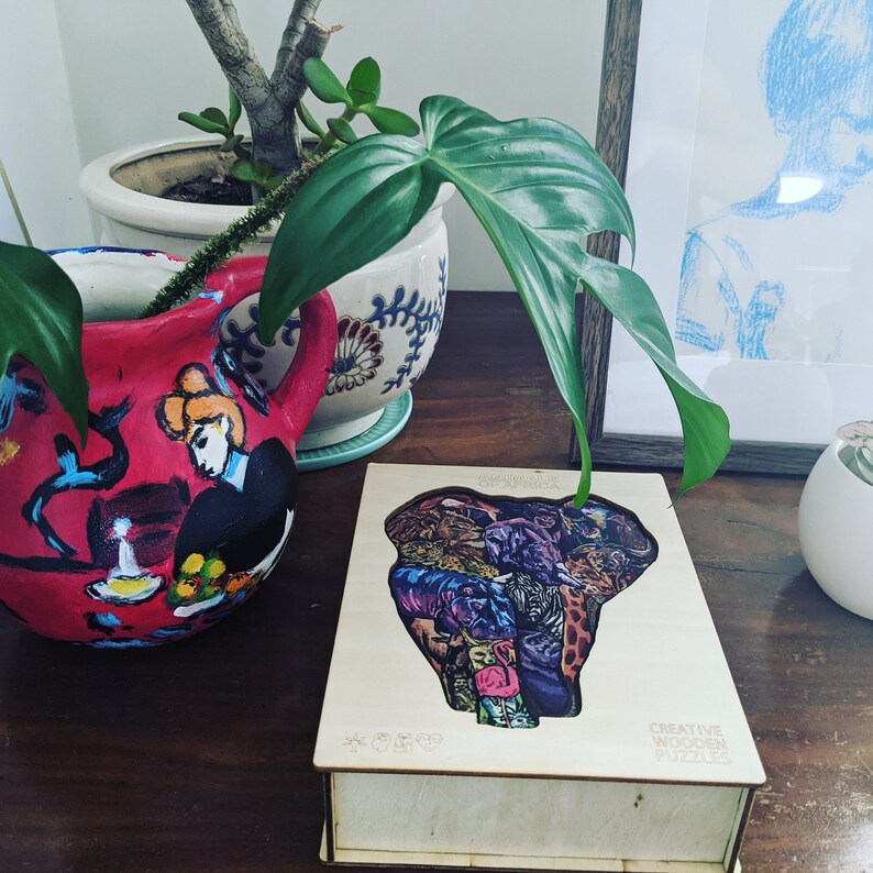 Puede incluir: Una caja de rompecabezas de madera con un dise&ntilde;o de elefante colorido. La caja est&aacute; abierta y las piezas del rompecabezas son visibles. La caja est&aacute; sobre una mesa de madera con una planta y un marco de imagen en el fondo.