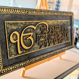 Puede incluir: Una placa de pared rectangular con un acabado desgastado en negro y dorado y un borde decorativo. La placa muestra el símbolo Gurmukhi Ik Onkar en oro y el texto Mool Mantar en letras doradas en relieve sobre un fondo oscuro texturizado.